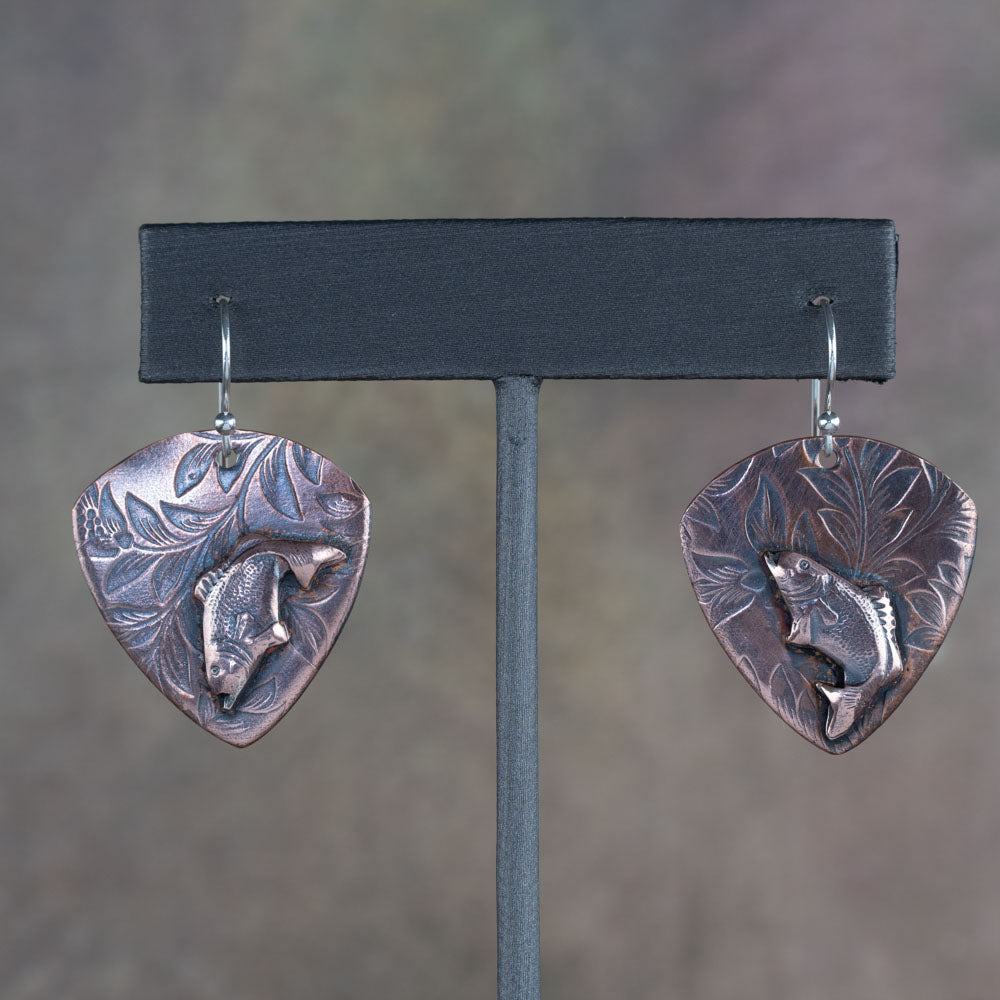 Handcrafted copper earrings featuring detailed jumping bass fish on a botanical-etched background – rustic nature-inspired fishing jewelry by Divella Designs, displayed on earthy surface.