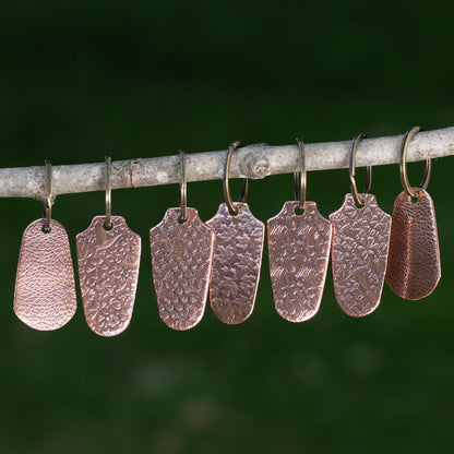 Handcrafted copper keychains with assorted embossed nature patterns hanging on a branch, artisan-made with rustic textures.