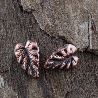 Handcrafted copper leaf post earrings with oxidized details, featuring realistic vein texture and asymmetrical heart-shaped foliage design on rustic wood background.