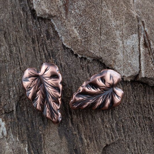Handcrafted copper leaf post earrings with oxidized details, featuring realistic vein texture and asymmetrical heart-shaped foliage design on rustic wood background.