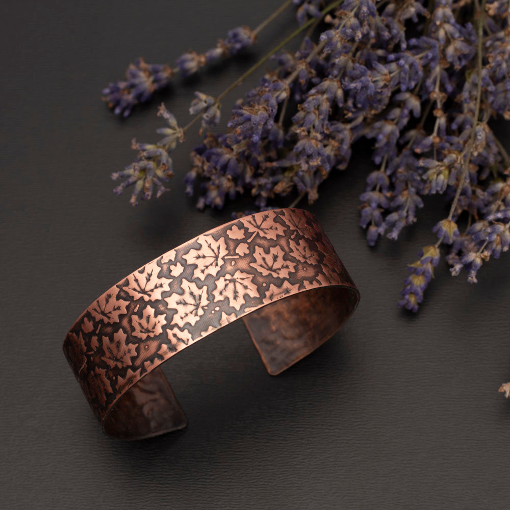 Etched copper cuff bracelet featuring a maple leaf pattern, displayed beside dried lavender on a dark background.