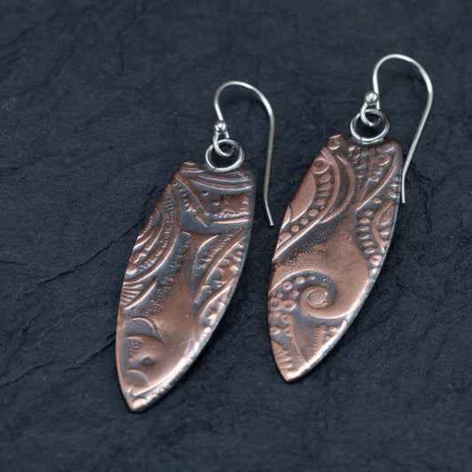 Handcrafted copper earrings with elongated surfboard shape and intricate embossed patterns, oxidized finish on sterling silver ear wires – artisan design by Divella Designs, displayed on black slate background.