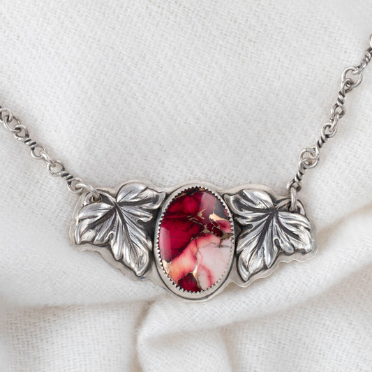 Close up of Crimson Bloom necklace with oval dahlia rose stone in pink and red, framed by two detailed sterling silver leaves on a handcrafted silver chain, botanical artisan jewelry.