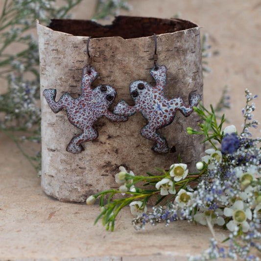 Whimsical handcrafted enamel earrings in the shape of dancing toads with speckled red and white patterns, black eyes, and oxidized copper ear wires, displayed on birch bark with white and lavender wildflowers – Dancing Toads by Divella Designs."
