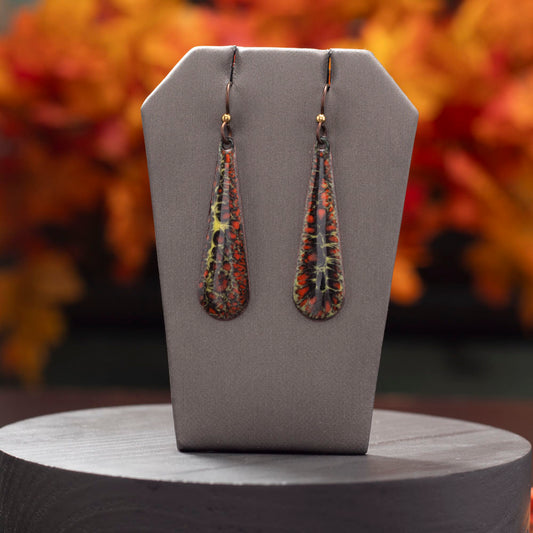 Dark orange and black enamel teardrop earrings with yellow veining, displayed on a gray stand with a warm, blurred autumn leaf background.
