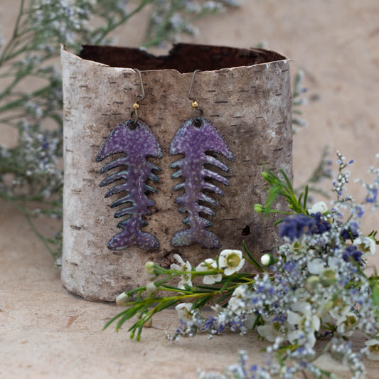 Handmade enamel earrings in a stylized fishbone design with a mottled purple and teal finish, accented with brass and copper ear wires, displayed on natural birch bark with delicate white and lavender flowers – Deep Current by Divella Designs.