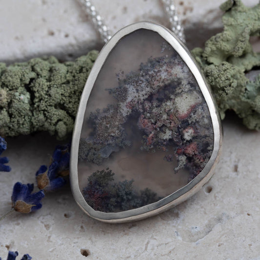 Teardrop-shaped pendant with dendritic agate stone on a textured surface with moss and flowers.