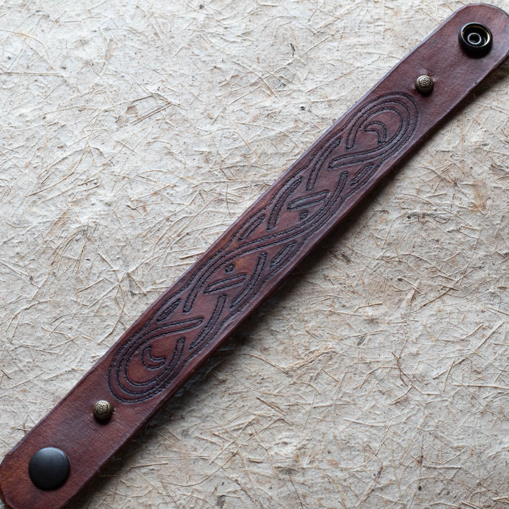 Brown leather bracelet with embossed design and metal fasteners on a textured beige background