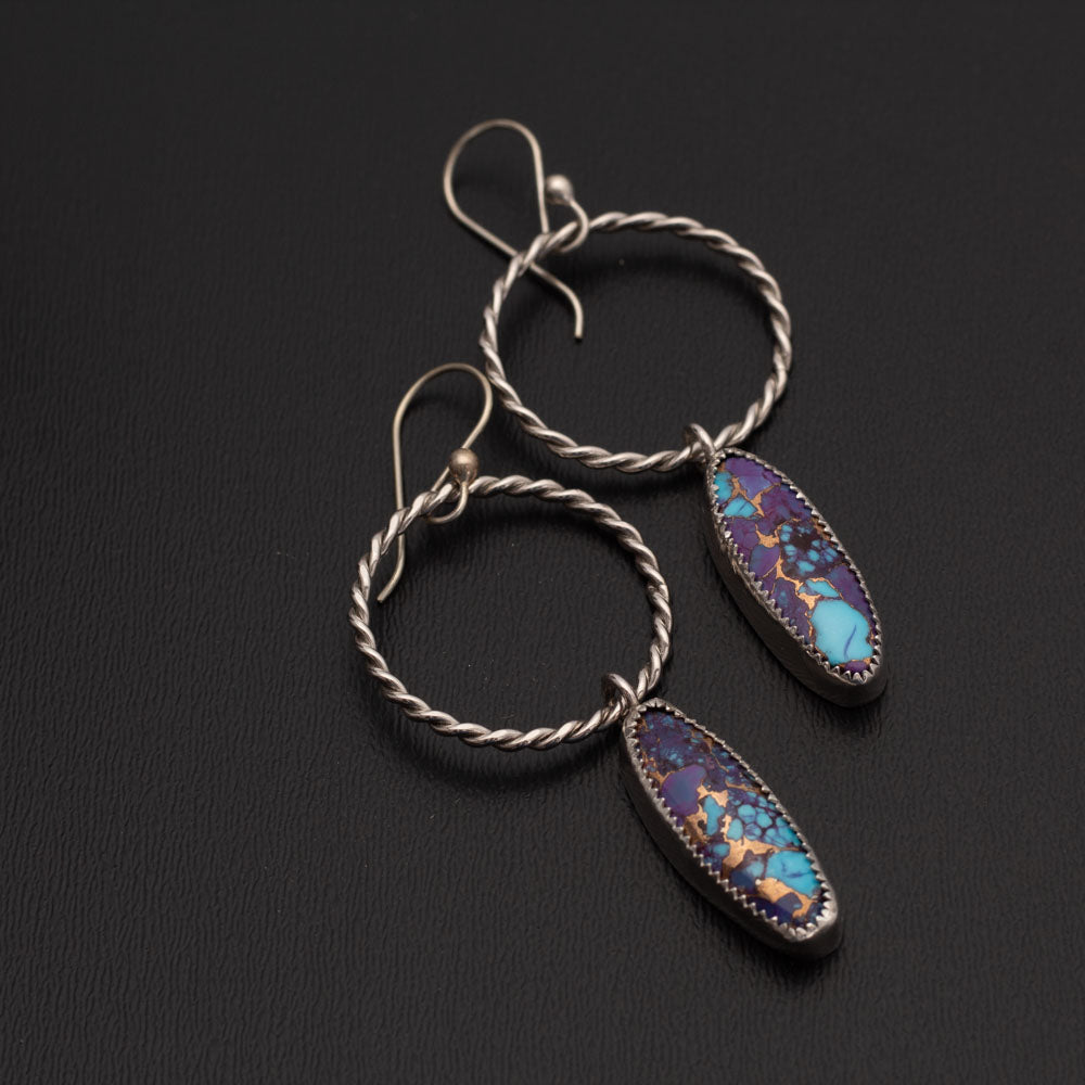 Handcrafted dangle earrings featuring oval purple Mohave turquoise and Kingman turquoise stones set in sterling silver with twisted silver hoops, displayed against a gray background.
