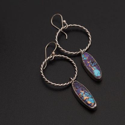 Handcrafted dangle earrings featuring oval purple Mohave turquoise and Kingman turquoise stones set in sterling silver with twisted silver hoops, displayed against a gray background.