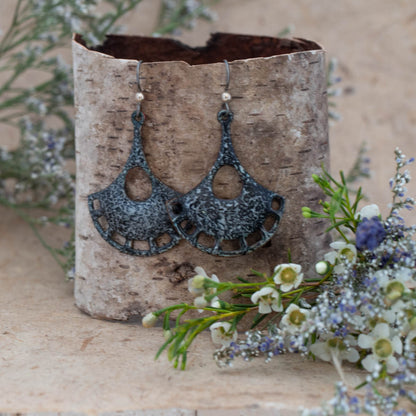 Handcrafted enamel earrings in a fan shape with black and gray speckled pattern. Artisan statement earrings with cut-out crescent design, displayed on birch bark with delicate wildflowers – Eclipse Fan Earrings by Divella Designs.