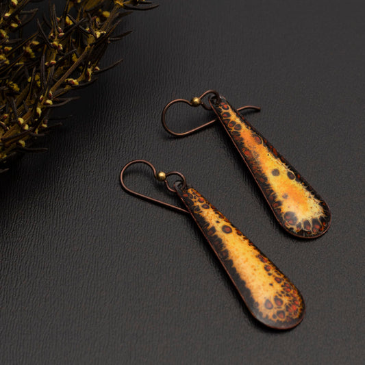 Long teardrop enamel earrings in warm tones of orange, yellow, and black with a speckled pattern, displayed on a dark textured background beside a sprig of dried foliage