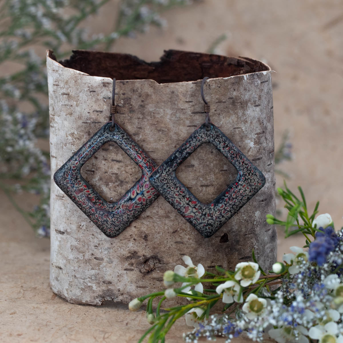Geometric square enamel earrings in a dark ember red and black lace pattern with open center. Handcrafted artisan jewelry by Divella Designs, displayed on natural birch bark with white wildflowers – Ember Lace Enamel Earrings.