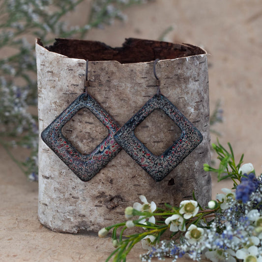 Geometric square enamel earrings in a dark ember red and black lace pattern with open center. Handcrafted artisan jewelry by Divella Designs, displayed on natural birch bark with white wildflowers – Ember Lace Enamel Earrings.