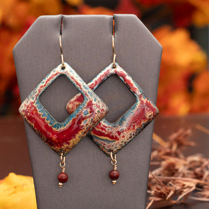 Square-shaped enamel earrings in fiery red, gold, and blue tones with center cut-outs and small red bead dangles, displayed on a dark surface with a fall leaf accents.