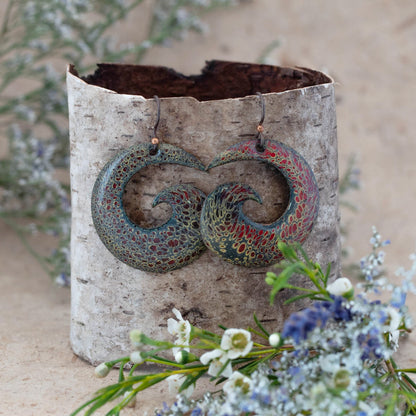 Artisan enamel earrings in a swirling wave design with fiery red, gold, and green tones, inspired by nature’s embers, displayed on birch bark with wildflowers – handmade by Divella Designs."