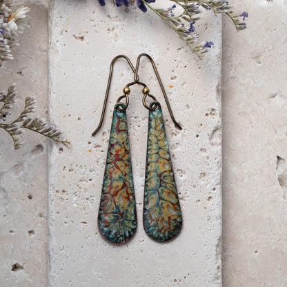 Handmade enamel teardrop earrings with earthy botanical pattern in yellow, teal, and rust tones, styled on natural stone with dried wildflowers – artisan jewelry by Divella Designs.