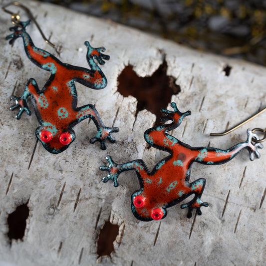 Handcrafted enameled copper frog earrings in bold red and teal with bright red eyes, tree frog design displayed on rustic birch bark – artisan nature jewelry by Divella Design
