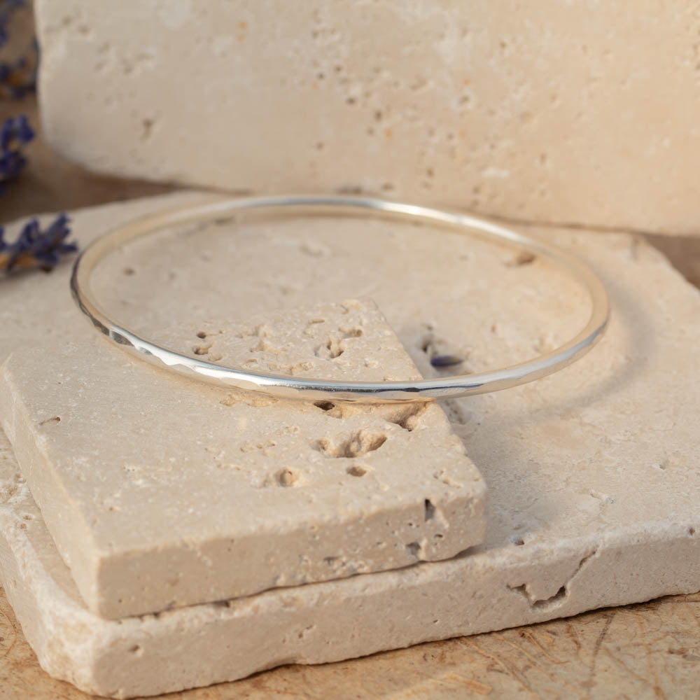 Handmade sterling silver faceted bangle with subtle hammered texture, photographed on stacked stone blocks with neutral tones and lavender in the background.