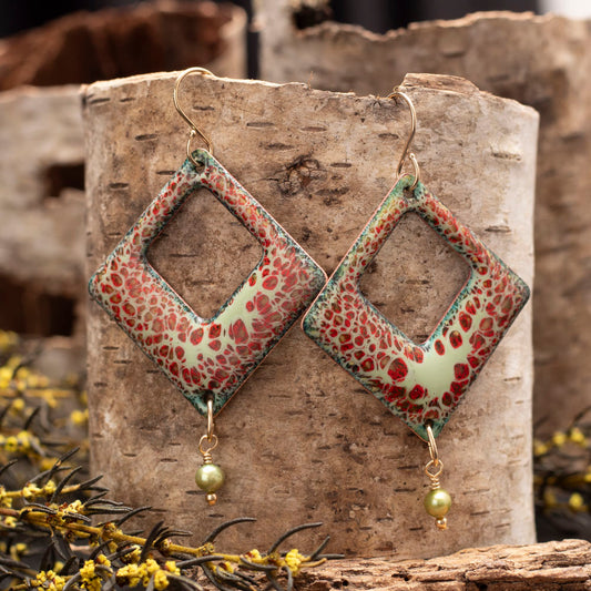 Square copper enamel earrings in mint green with red and black crackle design, finished with green pearl dangles and gold ear wires, styled on birch bark with yellow wildflowers.