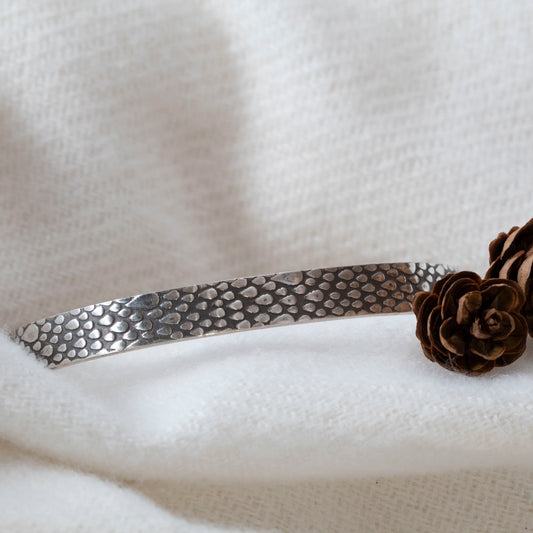 Sterling silver cuff bracelet with a raindrop texture, displayed on soft white fabric with small pinecones for decoration.