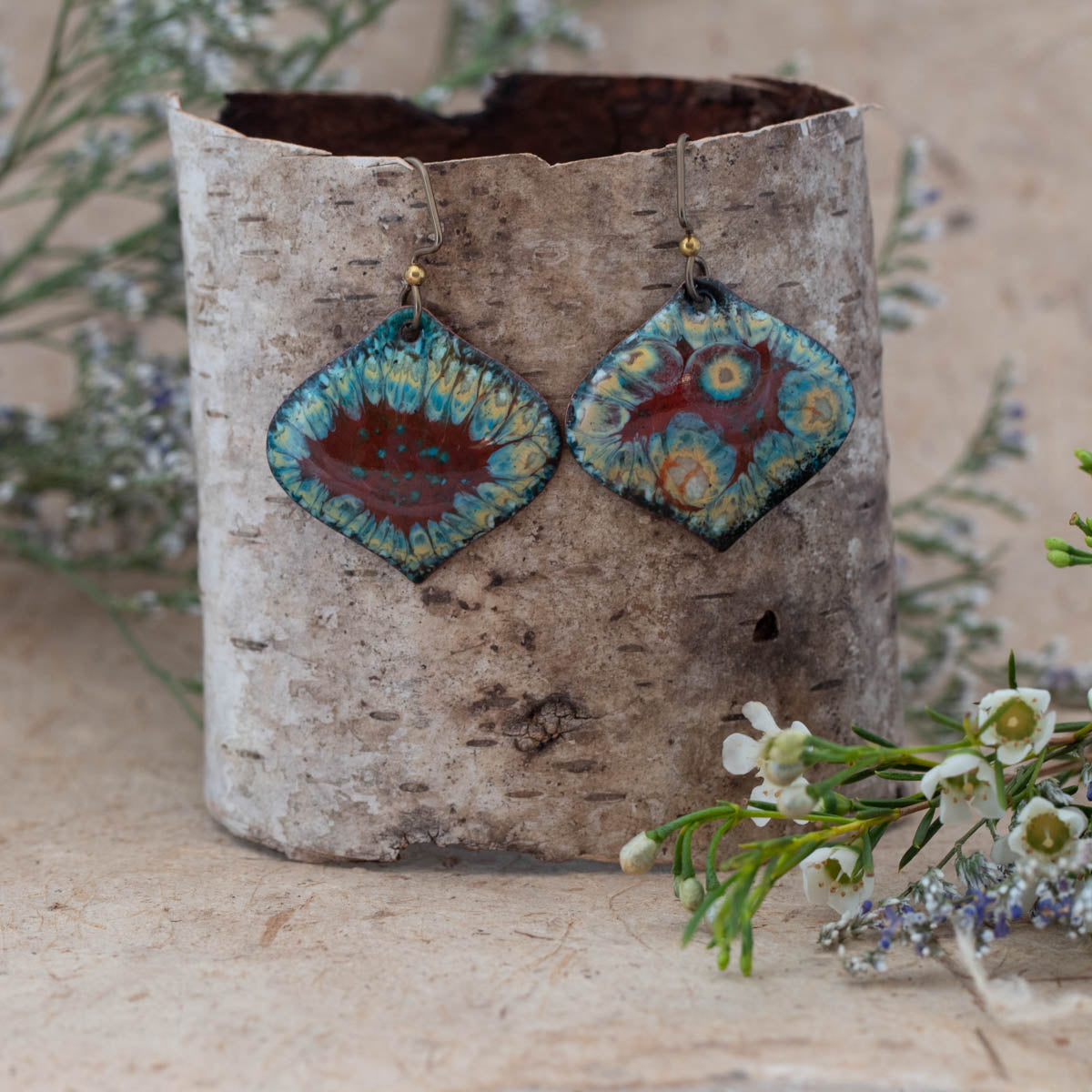 Handmade enamel earrings in a diamond shape with vibrant red, turquoise, and yellow floral-inspired patterns. Unique boho-style dangle earrings displayed on birch bark with wildflowers – artisan jewelry by Divella Designs.