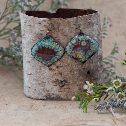 Handmade enamel earrings in a diamond shape with vibrant red, turquoise, and yellow floral-inspired patterns. Unique boho-style dangle earrings displayed on birch bark with wildflowers – artisan jewelry by Divella Designs.