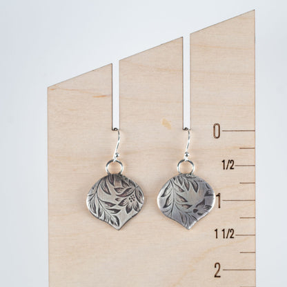 Handcrafted sterling silver earrings with etched botanical leaf and flower pattern, soft matte finish and organic shape – artisan nature-inspired jewelry by Divella Designs, shown on wooden ruler for scale.