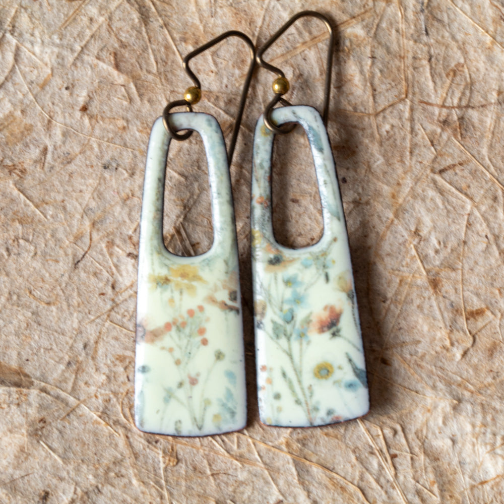 Floral enamel rectangular earrings with open cutouts, pale background, muted blue and yellow flowers, and niobium ear wires.