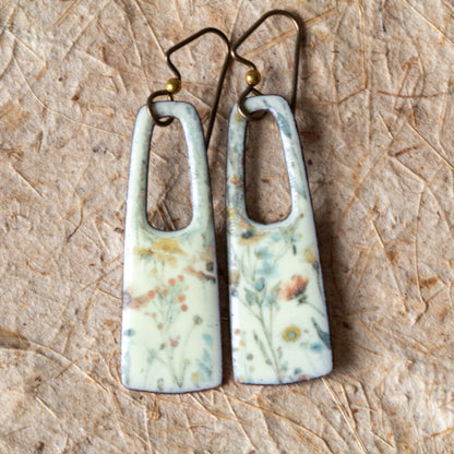 Floral enamel rectangular earrings with open cutouts, pale background, muted blue and yellow flowers, and niobium ear wires.