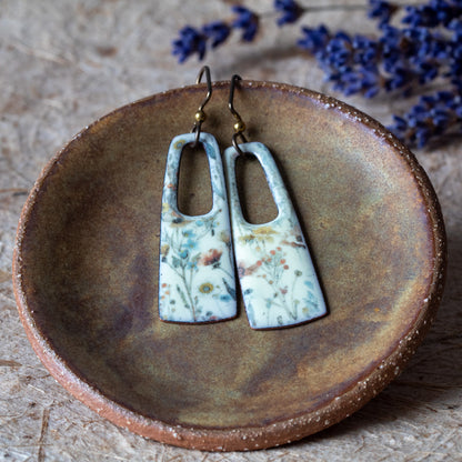 Floral enamel rectangular earrings with open cutouts, pale background, muted blue and yellow flowers, and niobium ear wires.