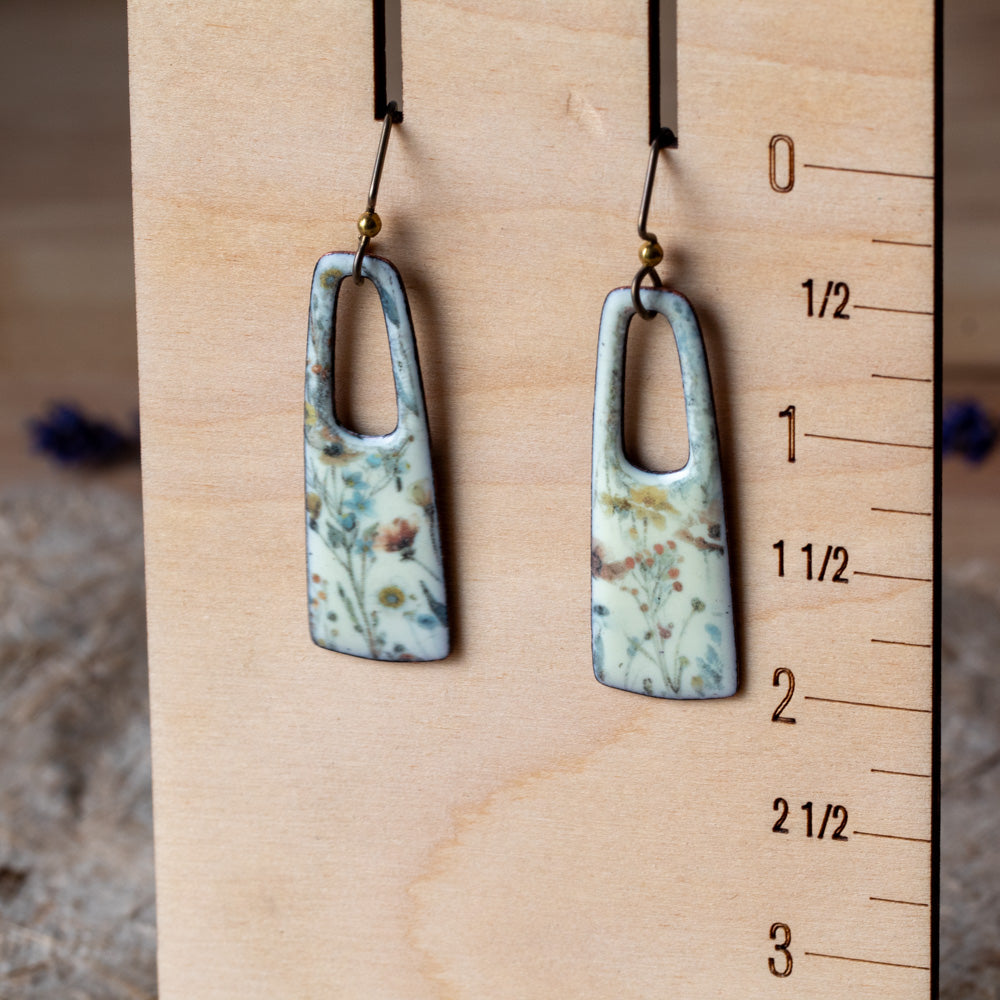 Floral enamel rectangular earrings with open cutouts, pale background, muted blue and yellow flowers, and niobium ear wires.