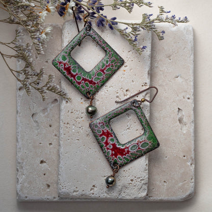 Handcrafted enamel earrings with red and green organic cell pattern, diamond shape with freshwater pearl dangles – Forest Cell design by Divella Designs, styled with dried florals on travertine tile.