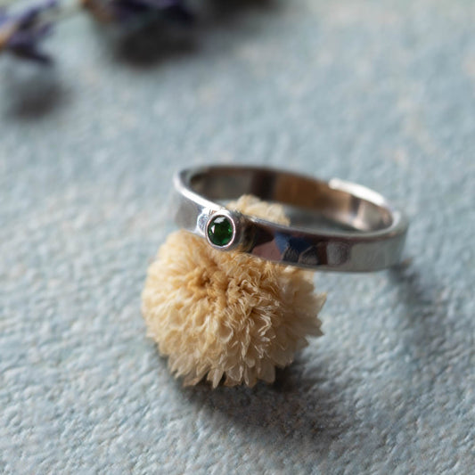 Handmade silver ring with small green stone in tube setting on soft blue surface with dried flower