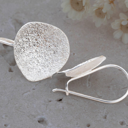 Silver textured round earring on a textured surface with flowers in the background