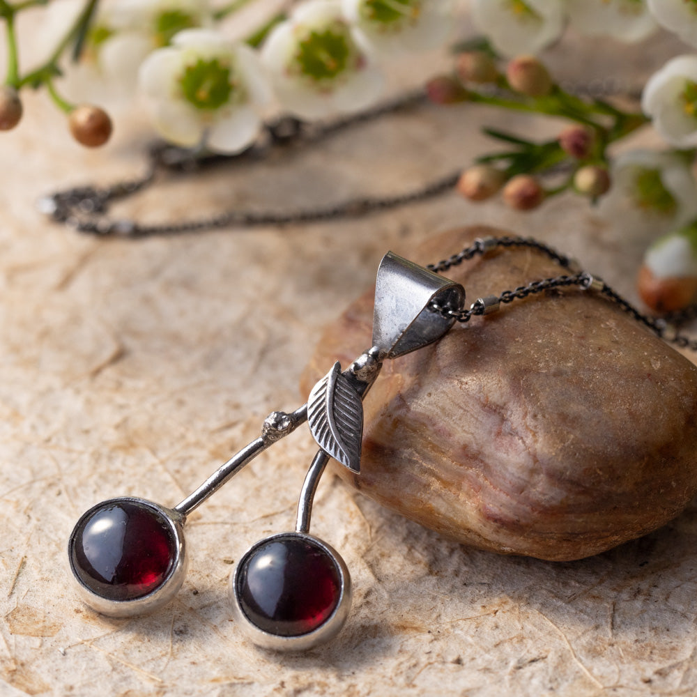 Garnet cherry pendant necklace handcrafted in fine and sterling silver resting against a natural stone with white flowers.