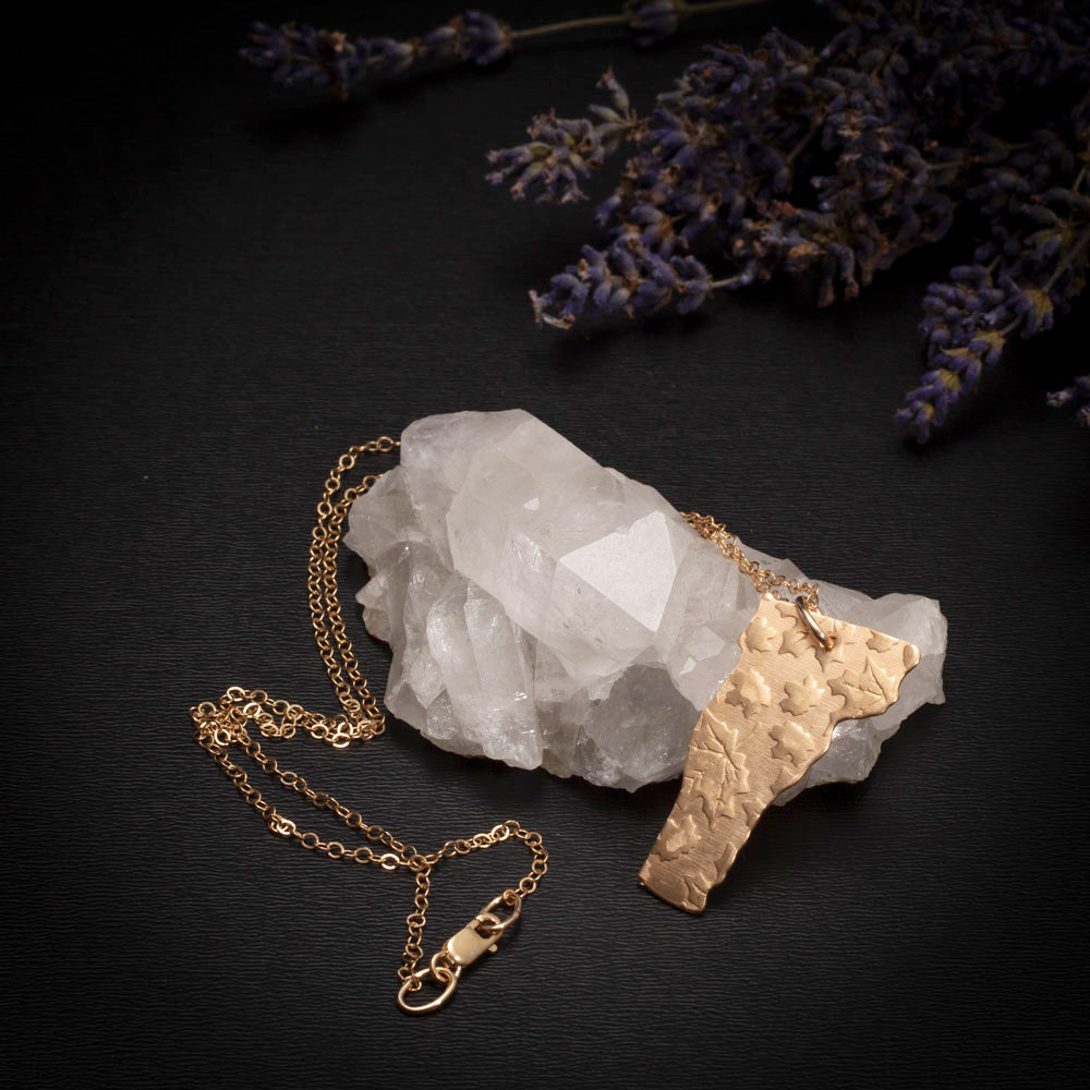 Brass Vermont-shaped pendant with embossed leaves on a gold-filled chain, displayed on a quartz crystal with lavender sprigs.