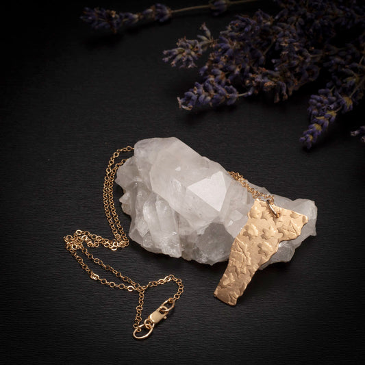 Brass Vermont-shaped pendant with embossed leaves on a gold-filled chain, displayed on a quartz crystal with lavender sprigs.