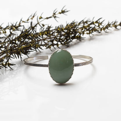 Handmade sterling silver cuff bracelet with oval green aventurine stone, minimalist artisan jewelry on white background with botanical accent.