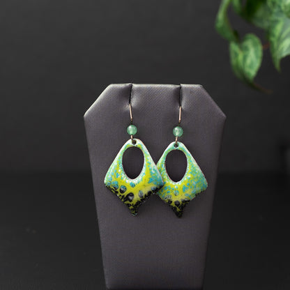 Handcrafted green and black enamel earrings with a diamond-shaped cutout design, textured lichen pattern, and niobium ear wires with green gemstone accents. Nature-inspired artisan jewelry.