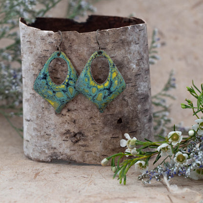 Handmade diamond-shaped enamel earrings with earthy green, yellow, and teal tones in a swirling pattern. Open oval center design with rustic patina, hanging on dark metal ear wires. Displayed on birch bark with wildflowers – Moonlight Earrings by Divella Designs.