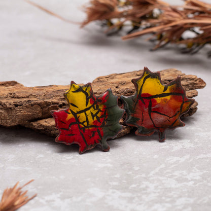 Colorful maple leaf-shaped earrings on a textured surface with dried plants in the background