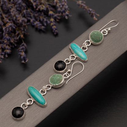 Handmade sterling silver earrings with turquoise, malachite, and onyx gemstones, arranged in a vertical drop design, displayed on a stand with lilacs in the background.