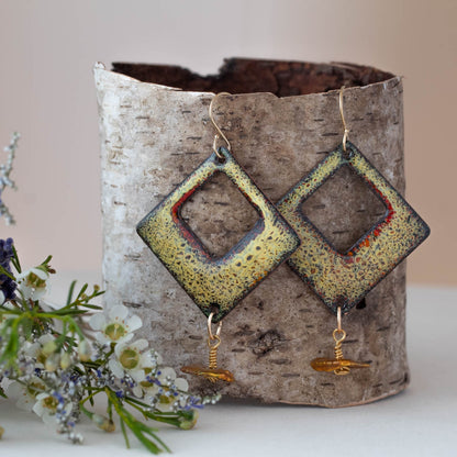 Diamond-shaped yellow enamel earrings with rustic red and black speckles, featuring dangling golden accent beads. Displayed on birch bark with white and purple flowers, handmade by Divella Designs.