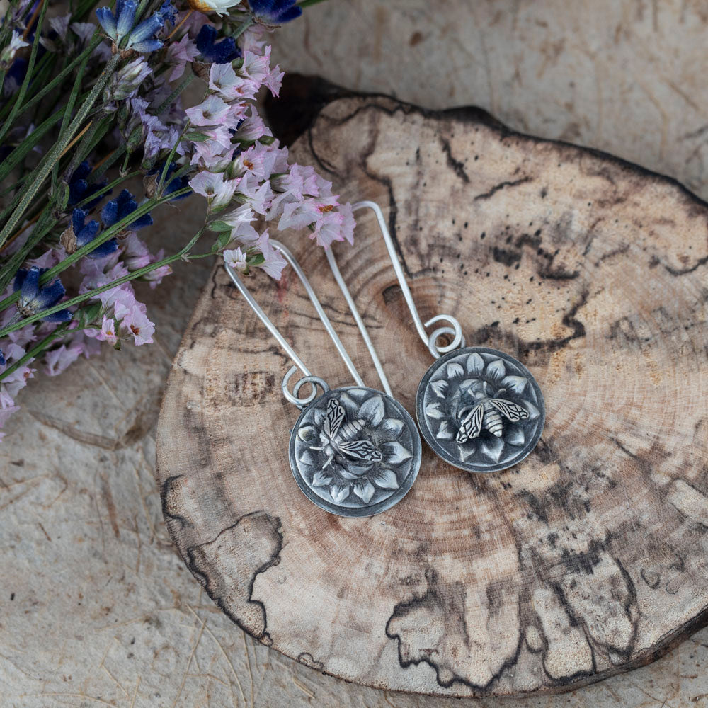 Sterling silver honeybee earrings by Divella Designs, handmade in Vermont with floral impressions and patinated satin finish, displayed on wood with dried wildflowers