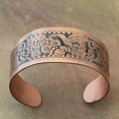 Copper bracelet with intricate floral and hummingbird design on a textured surface