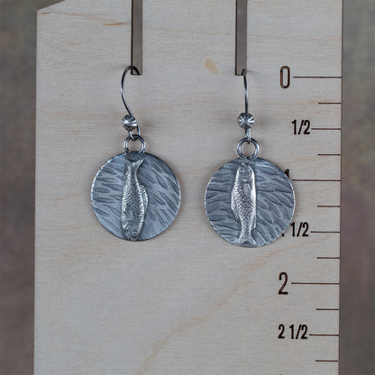 Handcrafted sterling silver earrings featuring detailed fish motifs swimming over textured water-like backgrounds – Swimmers artisan earrings by Divella Designs, displayed on a wooden ruler for scale