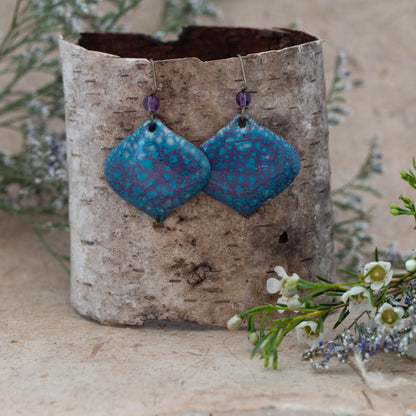 Indigo Bloom Enamel Earrings – vibrant handcrafted earrings in deep indigo and teal with petal-like patterns, paired with purple glass bead accents. Displayed on rustic birch bark with wildflowers.