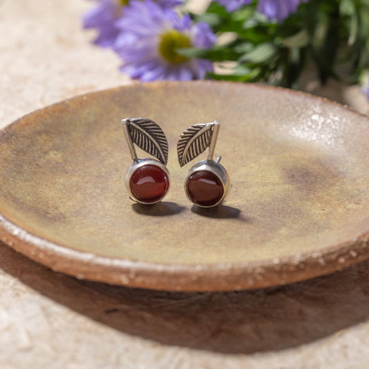 A pair of silver leaf stud earrings with round reddish-brown carnelian gemstones, displayed on a rustic ceramic plate with blurred purple flowers in the background.