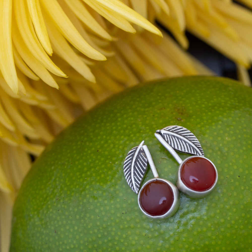 A pair of silver leaf stud earrings with carnelian gemstone centers, displayed on a green citrus fruit with blurred yellow flowers in the background.